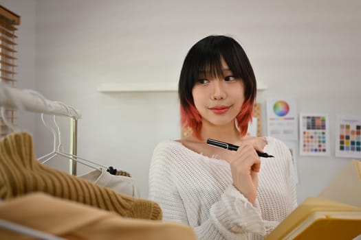 Photo of female fashion designer standing near clothing rack, working on new womenswear collection in modern studio.