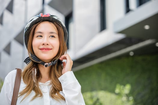 Close up smiling woman wearing helmet exercise outdoors with bicycle on street, Asian young businesswoman putting biking helmet for safety prepared cyclists around building go to work, Bike commuting