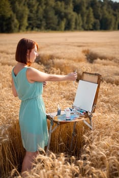 Woman with short red hair in blue dress starts painting in the field of ripe wheat, horizontal shot