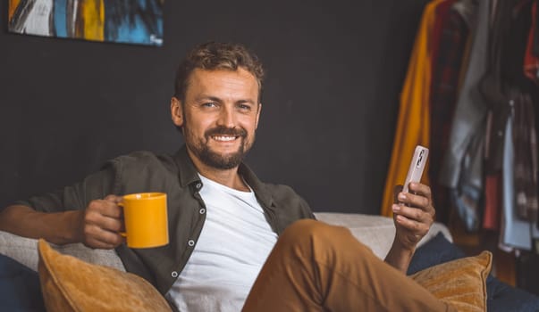 Happy man at home, enjoying cup of coffee while using phone. Concept of technology and communication is emphasized, highlighting ease and convenience of staying connected to others at any time. High quality photo