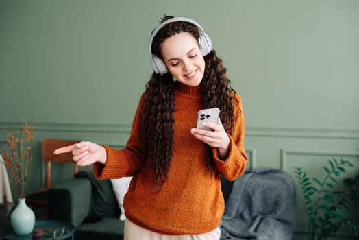 Happy pretty young woman wearing headphones using a smartphone dancing at home. Cheerful girl listening music on mobile phone, singing song feeling relaxed standing in modern living room interior