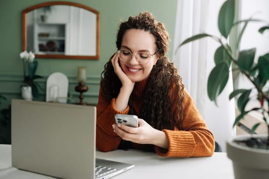 Young happy smiling pretty woman holding smartphone using cellphone modern technology, looking at mobile, checking cell phone apps, watching social media sitting at table at home