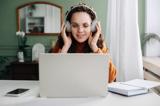 Young happy woman student wearing headphone using laptop elearning at home office listening music, live lecture or podcast webinar, learning language on online seminar sitting at desk with computer