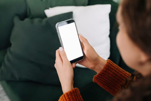 Over shoulder view of woman holding smartphone with white mock up cellular screen in hands using mobile apps on cell phone relaxing on sofa at home. Cellphone display mock up for advertising concept