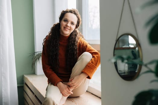 Pretty serene young woman sitting on windowsill relaxing at home looking at camera. Smiling calm lady chilling in apartment, dreaming, thinking of peaceful time enjoying peace of mind. Portrait.