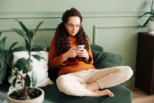 Relaxed young woman sitting on a sofa using a cell phone at home holding a smartphone, looking at cell phones shopping in mobile apps ordering e-commerce products, or checking social media