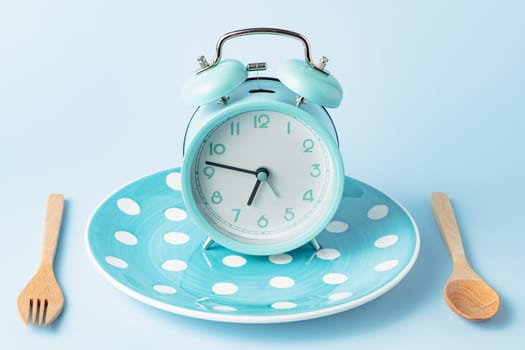 An alarm clock on an empty plate and cutlery set against blue background for the concept of food, time management, losing weight and eating on time.