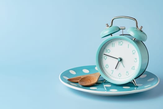 An alarm clock on an empty plate and cutlery set against blue background for the concept of food, time management, losing weight and eating on time.