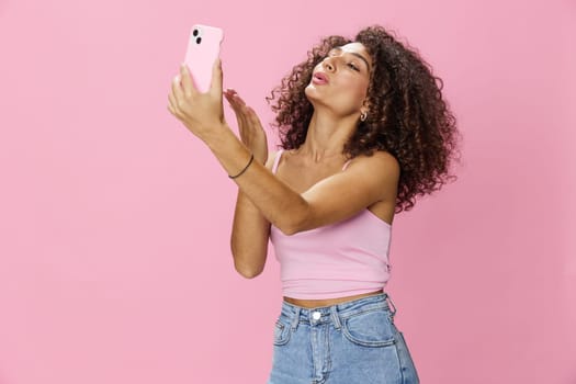 Woman blogger holding phone in hand video call, with curly hair in pink top and jeans poses on pink background, copy space, technology and social media, online. High quality photo
