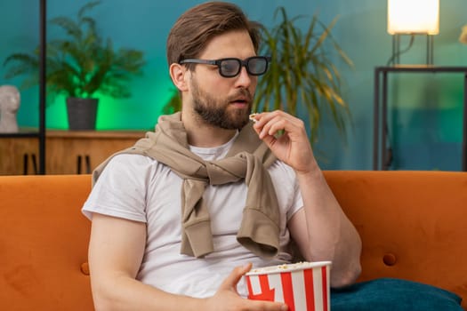Excited young man in 3D glasses sits on couch eating popcorn snacks and watching interesting TV serial sport game, film online social media movie content at home apartment. Guy enjoying entertainment