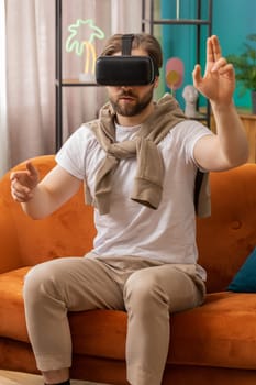Excited man using virtual reality futuristic technology VR app headset helmet to play simulation 3D 360 video game watching film movie. Portrait of guy in goggles at modern home apartment room on sofa
