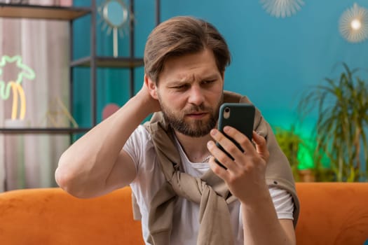 Sad displeased man use smartphone typing browsing, loses becoming surprised sudden lottery results, bad news, fortune loss, fail, deadline, virus. Young Caucasian one guy at home living room on couch