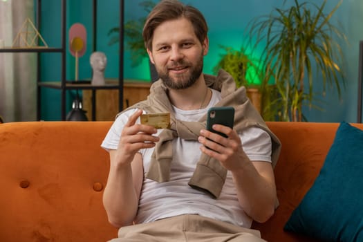 Caucasian man using credit bank card and smartphone while transferring money, purchases online shopping cashless order food delivery at home apartment indoors. Happy young guy sitting in room on couch