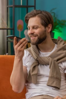 Happy excited young man making online phone call conversation with friends. Portrait of Caucasian guy enjoying mobile loudspeaker talking chatting communication at home apartment living room on couch