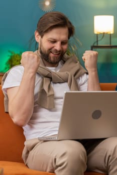 Surprised Caucasian man using laptop computer, receive mail good news message, shocked by sudden victory celebrate lottery jackpot win purchases online shopping at home apartment on sofa. Lifestyles