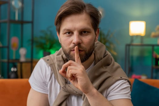 Shh be quiet, hush please. Caucasian man with presses index finger to lips makes silence hush gesture sign do not tells gossip secret. Young guy at modern home living room sitting on couch. Gossip