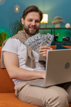 Planning budget. Rich happy Caucasian man counting money cash use laptop computer calculate domestic bills at home room. Young guy satisfied of income earnings, saves money for planned vacation, gifts