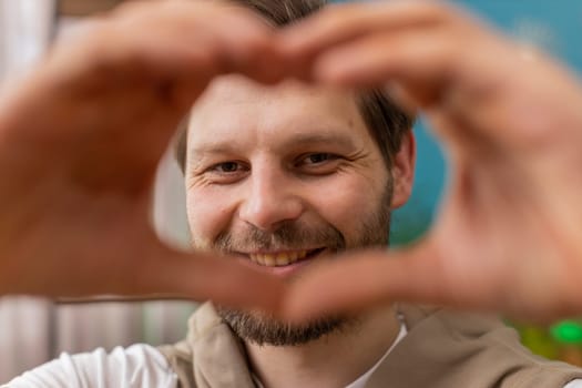 I love you. Happy Caucasian man at home living room couch makes symbol of love, showing heart sign to camera, express romantic feelings express sincere positive feelings. Charity, gratitude, donation