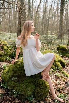 Snowdrops galanthus blonde. A girl in a white dress sits on a meadow with snowdrops in a spring forest.