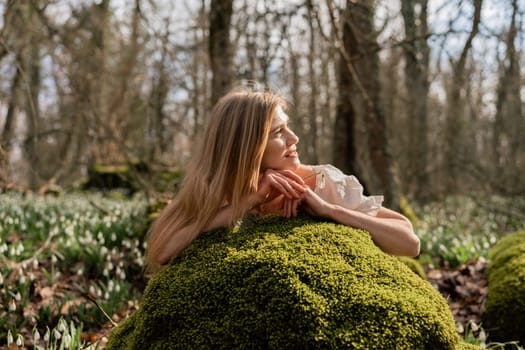 Snowdrops galanthus blond. A girl in a white dress lay down on a stone in the moss in a meadow with snowdrops in a spring forest.