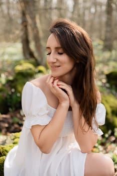 Portrait of a woman in the forest. She is sitting in a white dress on a meadow with snowdrops in a spring forest.