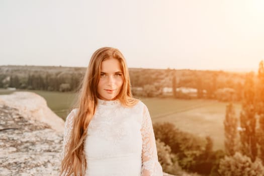 Romantic beautiful bride in white dress posing with sea and mountains in background. Stylish bride standing back on beautiful landscape of sea and mountains on sunset