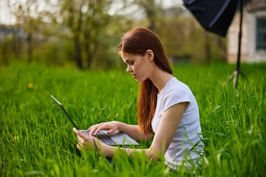 a woman in a light T-shirt sits in high green grass working at a laptop. High quality photo