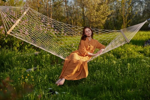 horizontal photo of a beautiful, red-haired woman lying in a hammock enjoying a rest in a long orange dress, on a warm summer day, smiling happily looking at the camera. High quality photo