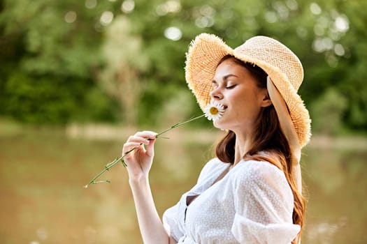 close-up portrait of a happy woman in a wicker hat with a camomile in her hands on the background of the lake. High quality photo