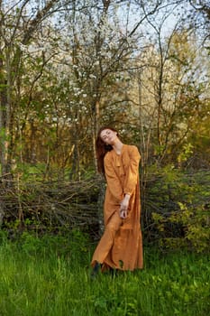 an elegant, sophisticated woman poses relaxed standing near a wicker fence at the dacha in a long orange dress, looking pleasantly at the camera. High quality photo