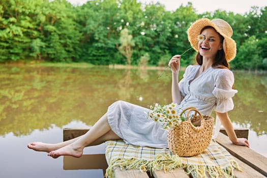 a happy woman with a chamomile flower in her hands sits on a pier by the lake and looks at the camera smiling. High quality photo