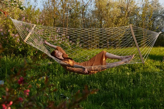 a happy woman in a long orange dress is relaxing in nature lying in a mesh hammock enjoying summer and vacation, looking at the sky. High quality photo