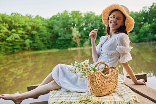 a happy woman with a chamomile flower in her hands sits on a pier by the lake and looks at the camera smiling. High quality photo