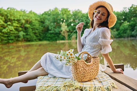 a happy woman with a chamomile flower in her hands sits on a pier by the lake and looks at the camera smiling. High quality photo