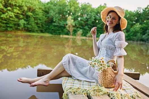 a happy woman with a chamomile flower in her hands sits on a pier by the lake and looks at the camera smiling. High quality photo