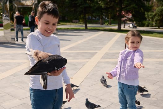 Adorable kids, brother and sister, feeding pigeons in the city square on a sunny day. The concept of kindness, care for animals.