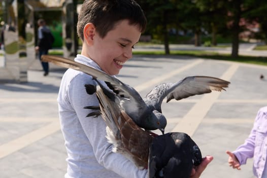 Portrait of a handsome happy cheerful child boy smiling, having fun feeding a flying flock of pigeons in summer park. The concept of happy carefree childhood and instilling love for nature and animals