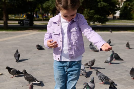 Friendly joyful child 5 years old, adorable little girl feeding pigeons in city summer park. The concept of kindness and care for animals. Childhood. Children. People. Nature