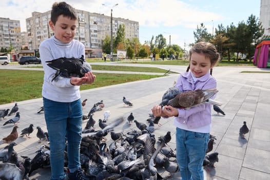 Two adorable kids, brother and sister feed pigeons in the park square. Spring. Lifestyle. The concept of kindness, care for animals. Family outing. A walk with benefit. Children, nature and animals.