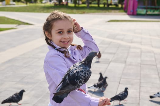 Lifestyle portrait of happy joyful lovely Caucasian child girl 5 years old, smiling cutely with beautiful toothy smile, looking at camera while feeding a pigeon sitting on her arm in city park square