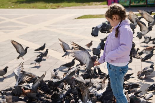 Adorable little child girl 5 years old, feeds flock of flying pigeons and birds in town square. Carefree childhood. Kids and nature. The concept of compassion, love, care and kindness for wild animals