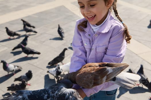 Selective focus. Overjoyed excited Caucasian little kid girl, smiling cheerfully, feeding pigeons outdoors. Lovely daughter expresing positive emotions, feeling connection with nature and wild animals