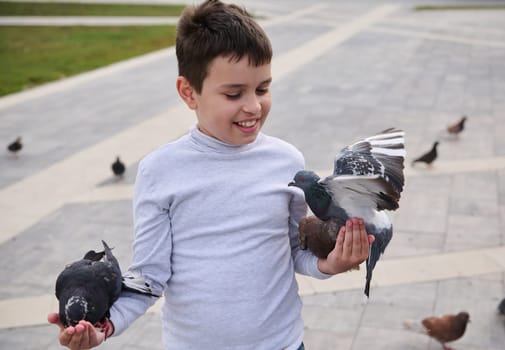 Caucasian adorable happy child boy, smiling, having fun, connecting with wild animals while feeding feral pigeons in the square of a city park. Children, nature, kindness and care for animals concept