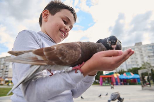 Adorable boy feeding rock doves sitting on his hands, smiling, expressing positive emotions while connecting with animals leaving on the street. The concept of childhood, care and love for animals