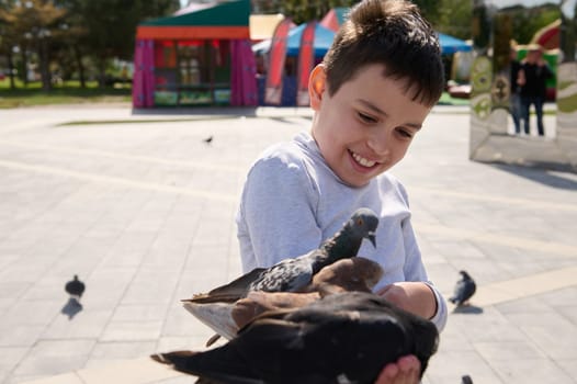 Friendly joyful pre teen boy, adorable child feeds pigeons in city summer park. The concept of kindness and care for animals. Childhood. Children. People. Nature