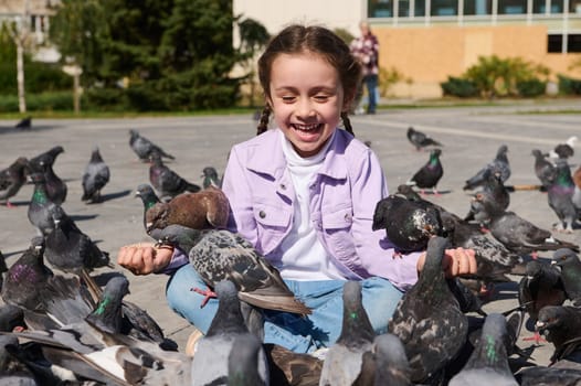 Adorable little Caucasian child girl sitting in lotus pose, feeding flock of flying pigeons and birds in town square. Carefree childhood. Kids and nature. The concept of care and kindness for animals