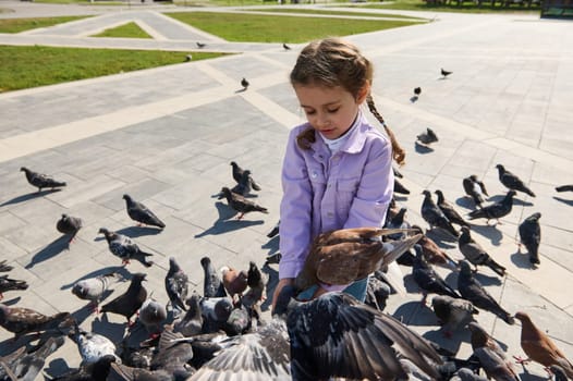 Adorable little kid girl connects with wild animals by feeding a flock of flying hungry pigeons in the city square. The concept of a happy carefree childhood and instilling love for nature and animals