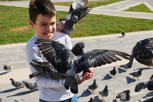 Caucasian teen boy feeding flock of flying pigeons in the summer city park square. The concept of care for wild animals. Wildlife and people. Children and nature