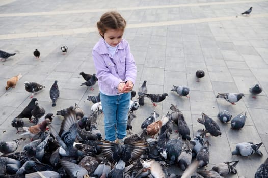 Caucasian adorable little girl, smiling, having fun, connecting with wild animals while feeding feral pigeons in the square of a city park. Children, nature, kindness and care for animals concept