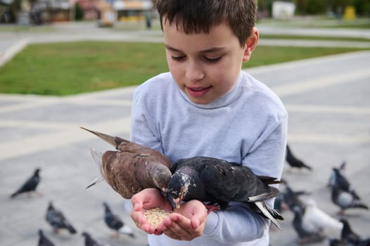 Close-up portrait of Caucasian adorable teen boy having fun during family outing, feeding birds from his hands in park square. The concept of kindness, walking with benefits, love and care for animals
